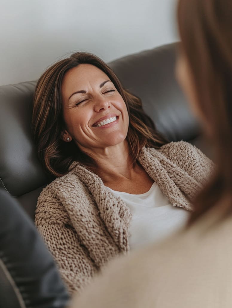 A woman with brown hair is leaning back on a dark sofa, smiling with her eyes closed. She is wearing a beige knit cardigan over a white top. Another person, slightly out of focus and with brown hair, appears in the foreground—perhaps they're discussing their experience at the Melbourne Counselling Centre.