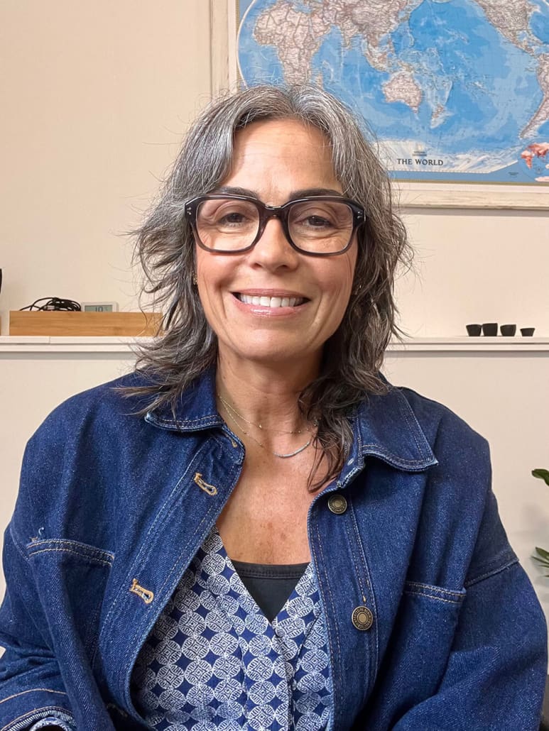 A person with gray hair and glasses smiles at the camera. They are wearing a blue denim jacket and a patterned shirt. A world map is visible on the wall behind them.