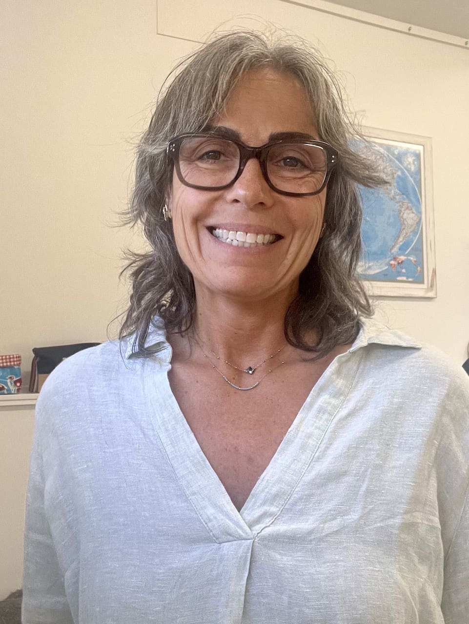 A person with shoulder-length hair and glasses smiles at the camera, wearing a light-colored button-up shirt. There is a wall with a framed picture in the background.